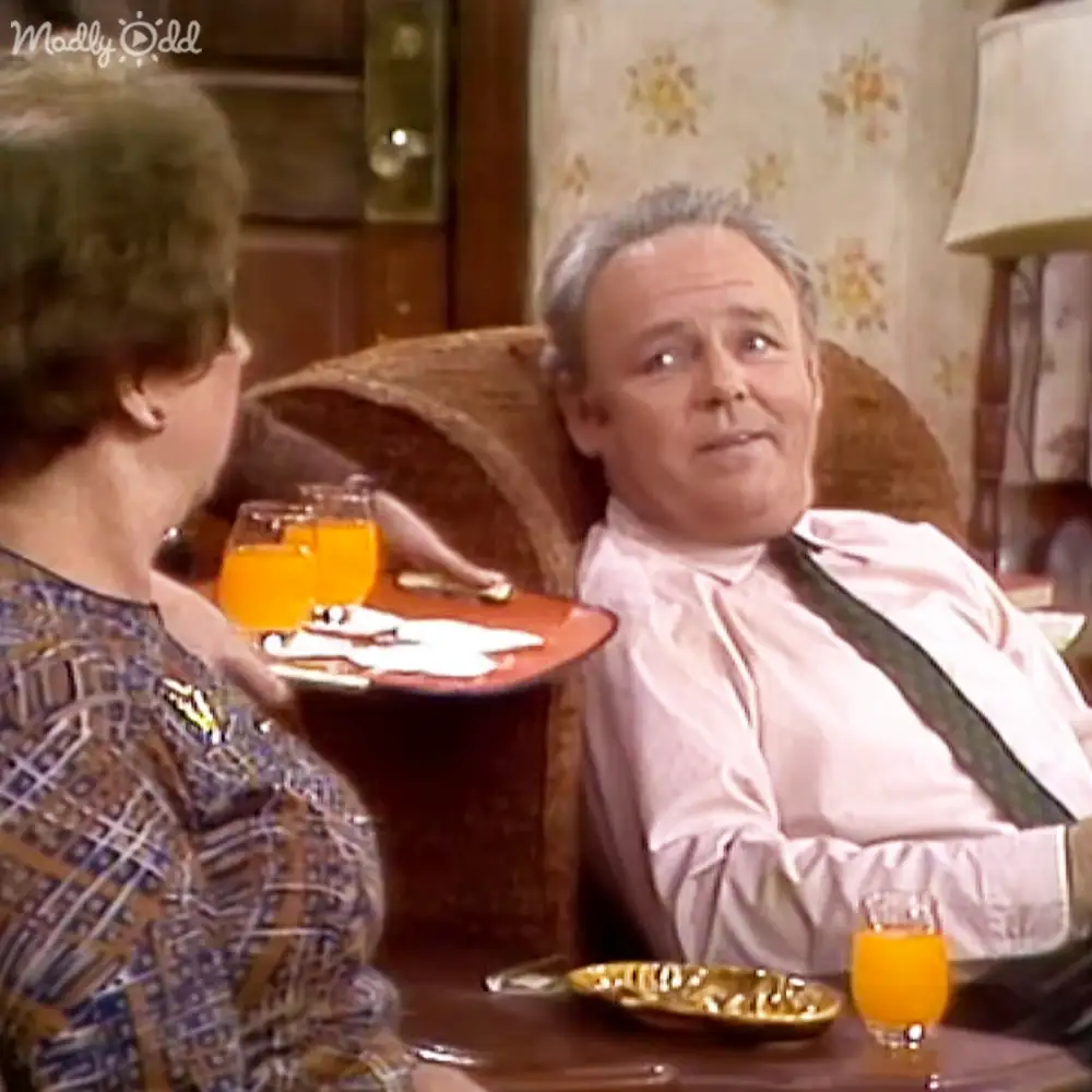 Middle-aged female in a patterned blouse, older male in a light pink dress shirt with dark suspenders, wicker chair and lamp visible in an indoor setting with floral wallpaper.