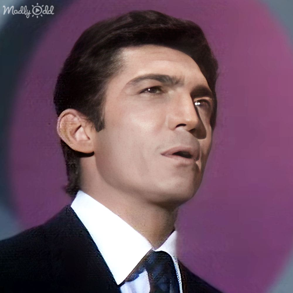 Young adult male wearing a dark suit, white dress shirt, and patterned tie in an indoor setting with purple hues.