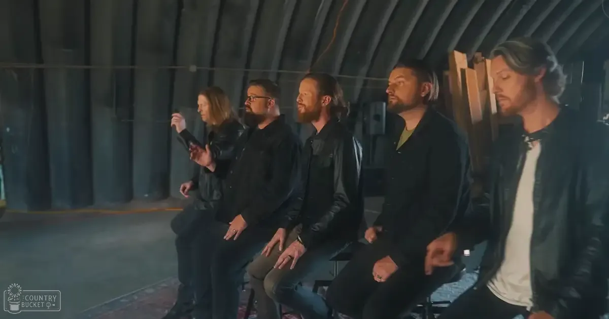 Group of adult males seated indoors against an industrial backdrop, wearing dark pants and black leather jackets over various shirts; natural light suggests daytime setting.