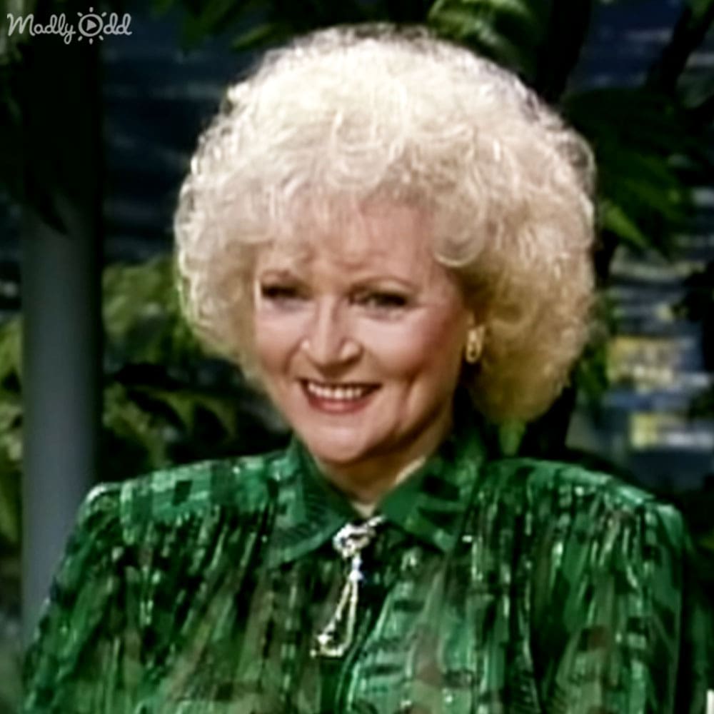 An older adult woman wearing a green patterned blouse, set against an indoor studio backdrop with plants and artificial lighting.