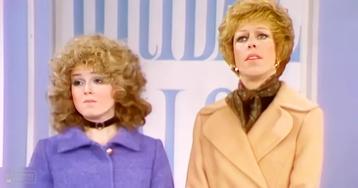 Two women in a studio setting, one wearing a purple coat with a high collar and red button, the other in a beige coat over patterned scarf, both with gold earrings; backdrop suggests professional lighting.