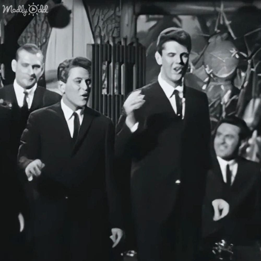 A young adult male stands at center, dressed in a dark suit and white shirt with a dark tie, surrounded by other individuals similarly attired; setting appears to be an indoor stage or studio.