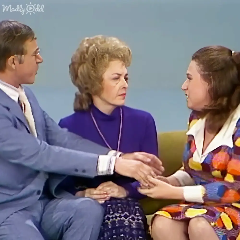 A studio setting with three individuals seated against a plain light blue wall, wearing formal and semi-formal attire.