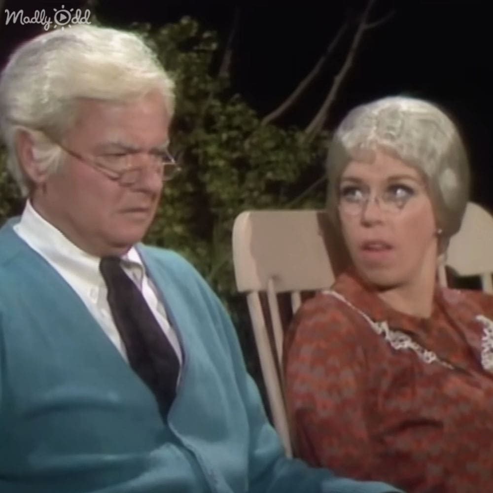 An older adult male in a light blue cardigan and an adult female in a patterned red dress at night outdoors, surrounded by trees with green foliage.