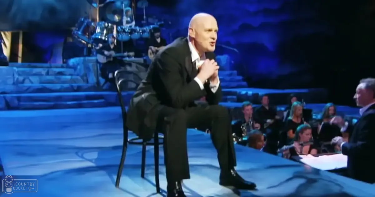 A middle-aged male singer performing on stage in a concert hall with an orchestra behind him.