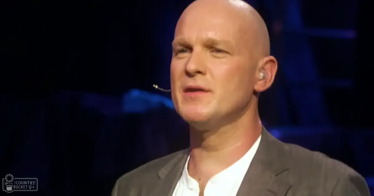 A middle-aged man speaks at a formal indoor event, wearing a dark gray suit jacket and white collared shirt with stage lighting behind him.