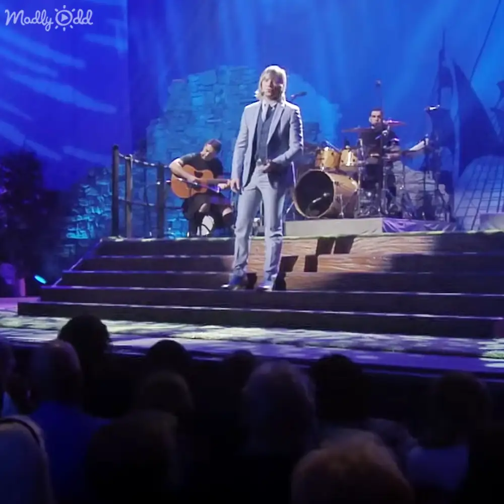 An adult male performer on stage at an indoor concert venue with blue lighting, surrounded by other musicians and audience members facing a stage with steps and rock-like structures.