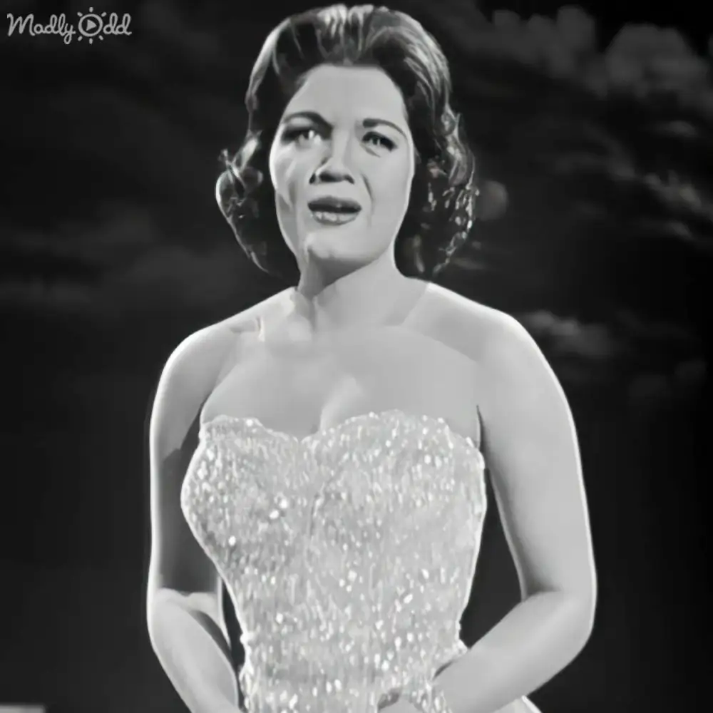 An adult female wearing a strapless, embellished gown with a fitted bodice and flared skirt, likely from the mid-20th century, indoors at what appears to be a formal event or performance setting.