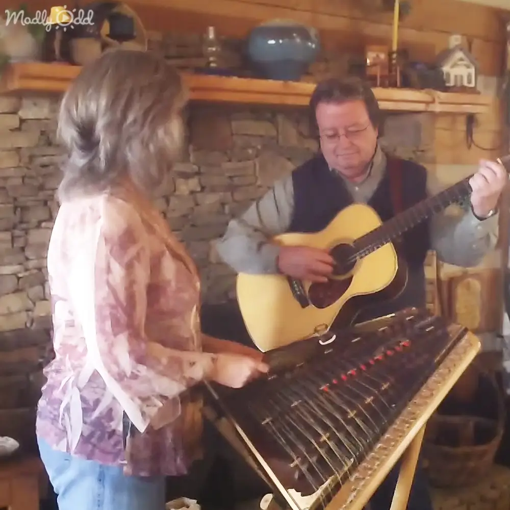 A rustic indoor setting with a musical performance; an adult female in a patterned top plays alongside an older male musician playing a psaltery, surrounded by stringed instruments and rustic decor.