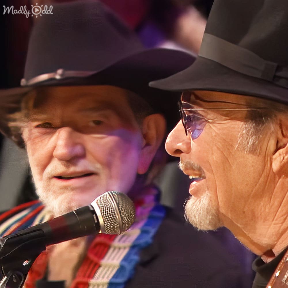An older adult male singer performing with another older adult male in a dimly lit indoor setting, likely part of a live performance or recording session.
