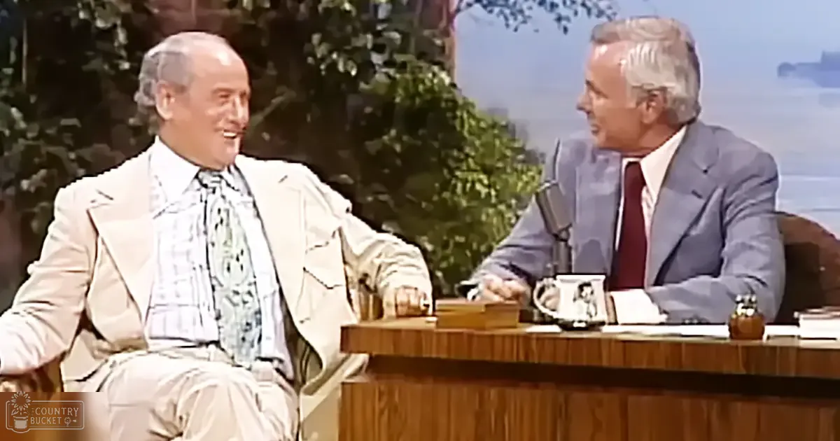 Two older adult men in formal suits seated at a desk outdoors, with trees and blurred cityscape in the background during spring or summer.