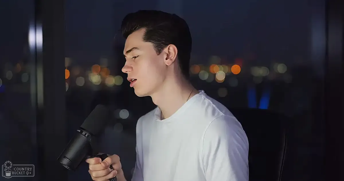 Young adult male wearing a white, relaxed-fit t-shirt with short sleeves, holding a black microphone indoors at night.