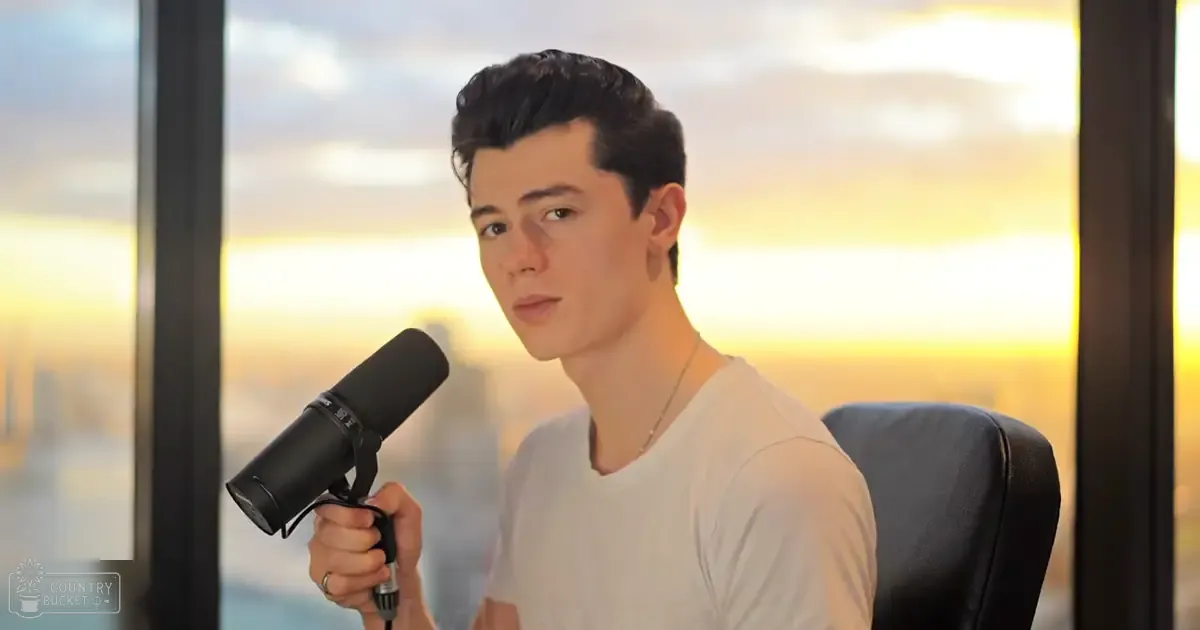 Young adult male in relaxed fit white t-shirt holding microphone against large window with sunset view.