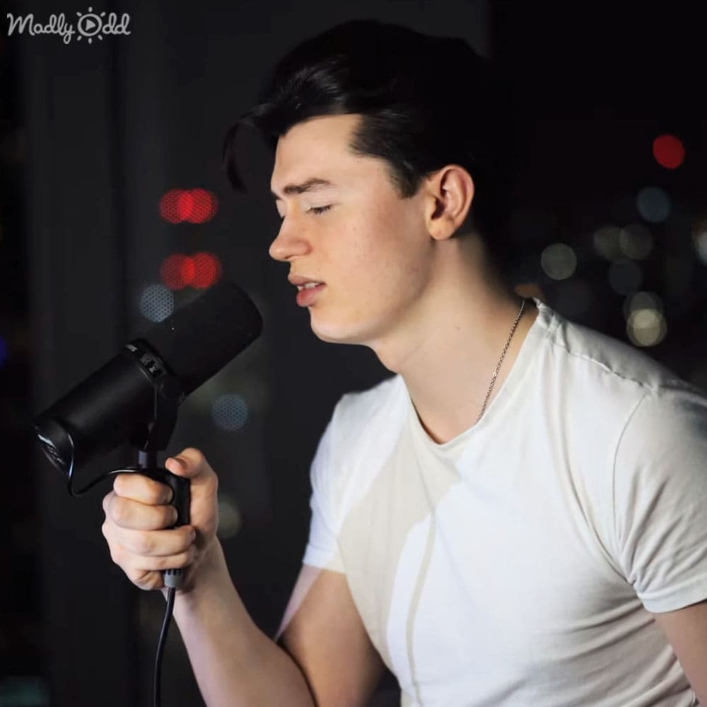 A young adult male wearing a relaxed, modern casual white t-shirt with cotton fabric, holding a microphone with a pop filter and wearing a thin chain necklace in an indoor setting with blurred city lights at night.