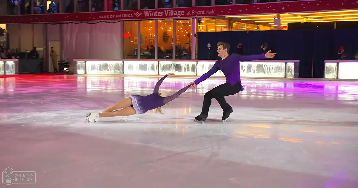 A young adult female figure skater in a purple dress at Bryant Park's Winter Village, surrounded by festive decorations under evening lights.