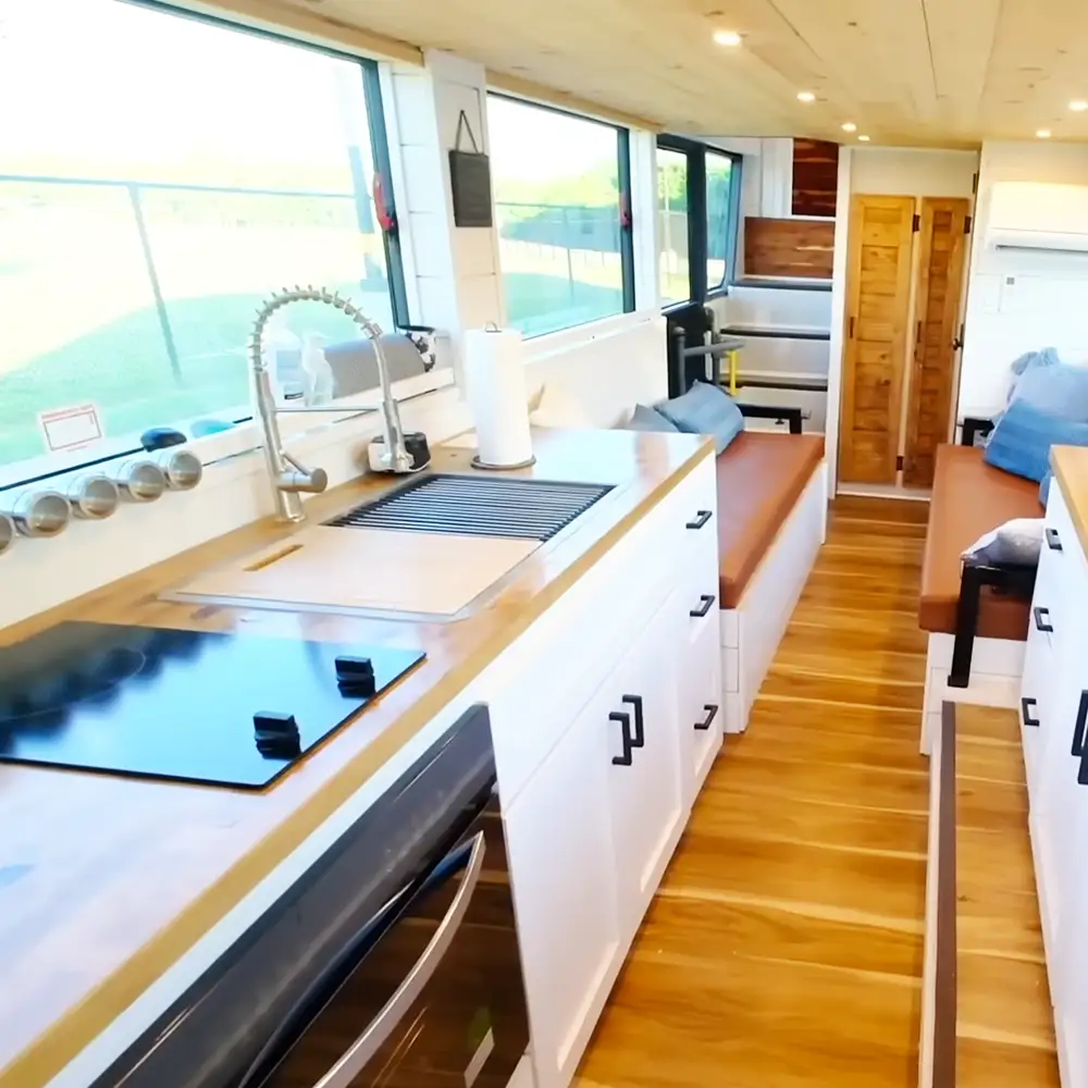 A modern kitchen area with white cabinets, black handles, a built-in cooktop, sink with faucet and cutting board, wooden flooring, bench seating near back wall, door to another room, blue cushions in background, natural light from large windows on daytime ambiance, dominant colors: white, brown, black, blue, green