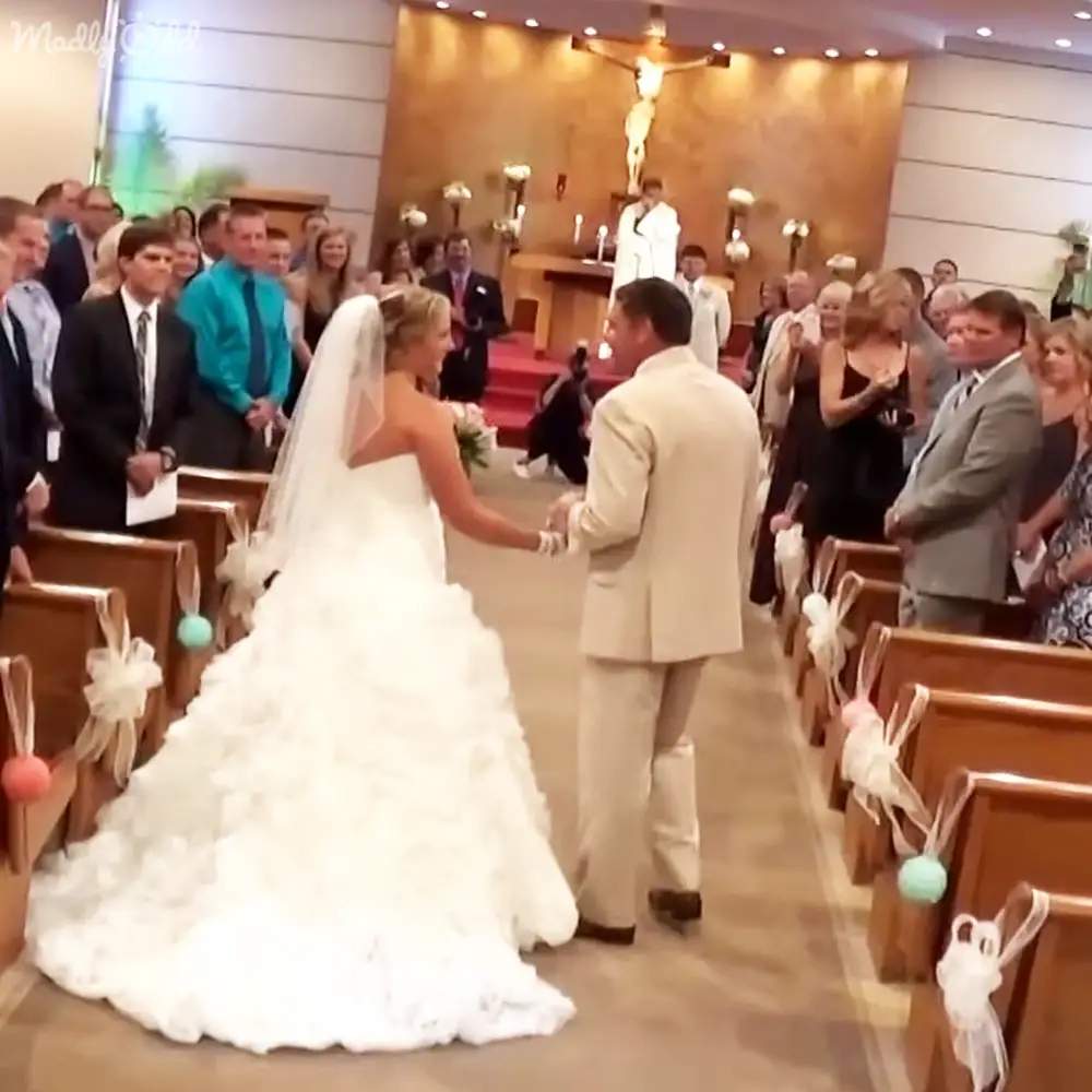 An adult female bride in a white strapless gown with an adult male groom in a light beige suit at an indoor church wedding ceremony, decorated pews and ribbons, bright even lighting from overhead lights.