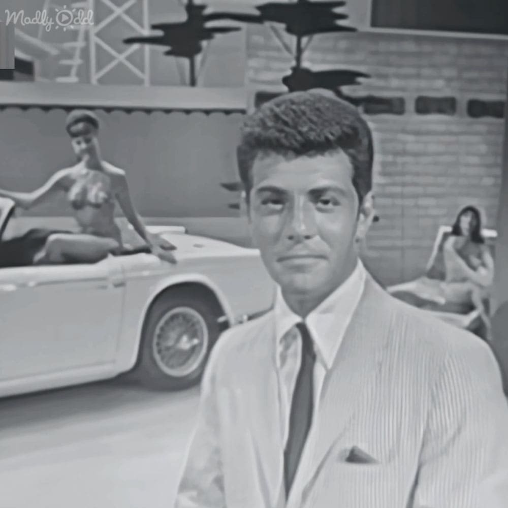 Urban setting with brick buildings, a white convertible car in the background, and individuals dressed in mid-20th century attire.
