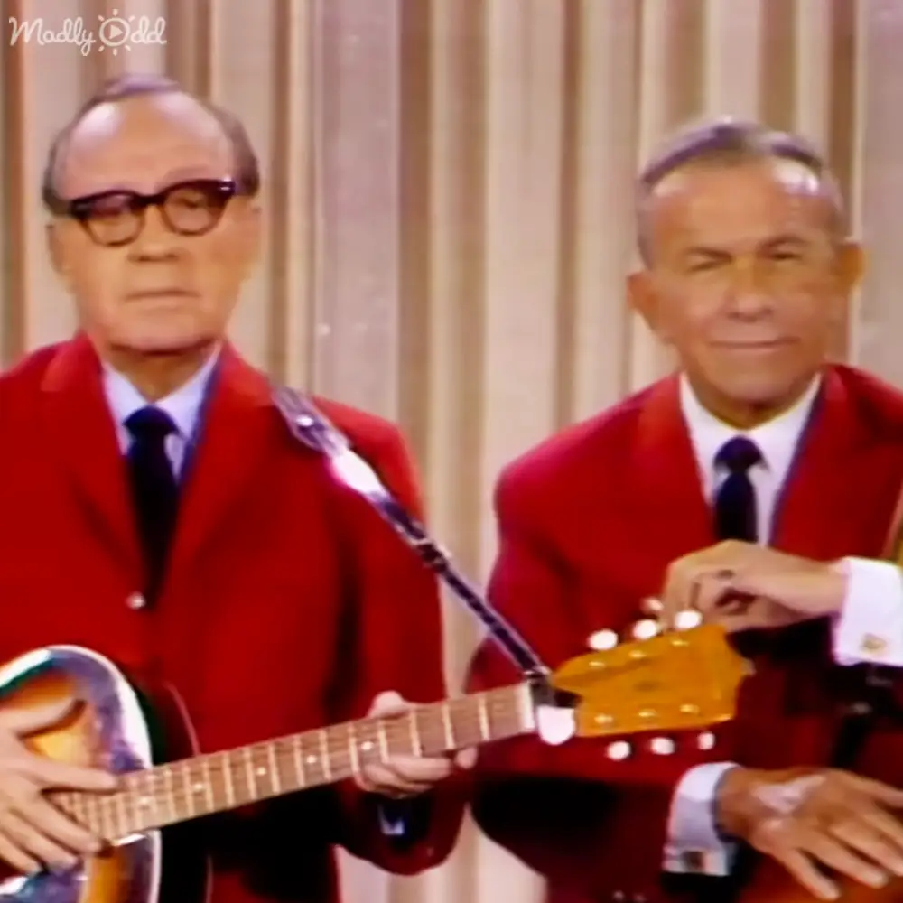 Musical performance scene with older adult male singers in red blazers, white shirts, and dark ties against beige curtains indoors under bright even lighting.