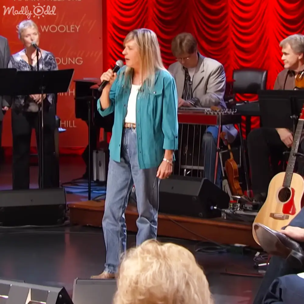 A middle-aged woman singing in a theater setting with red curtains and musical instruments visible.