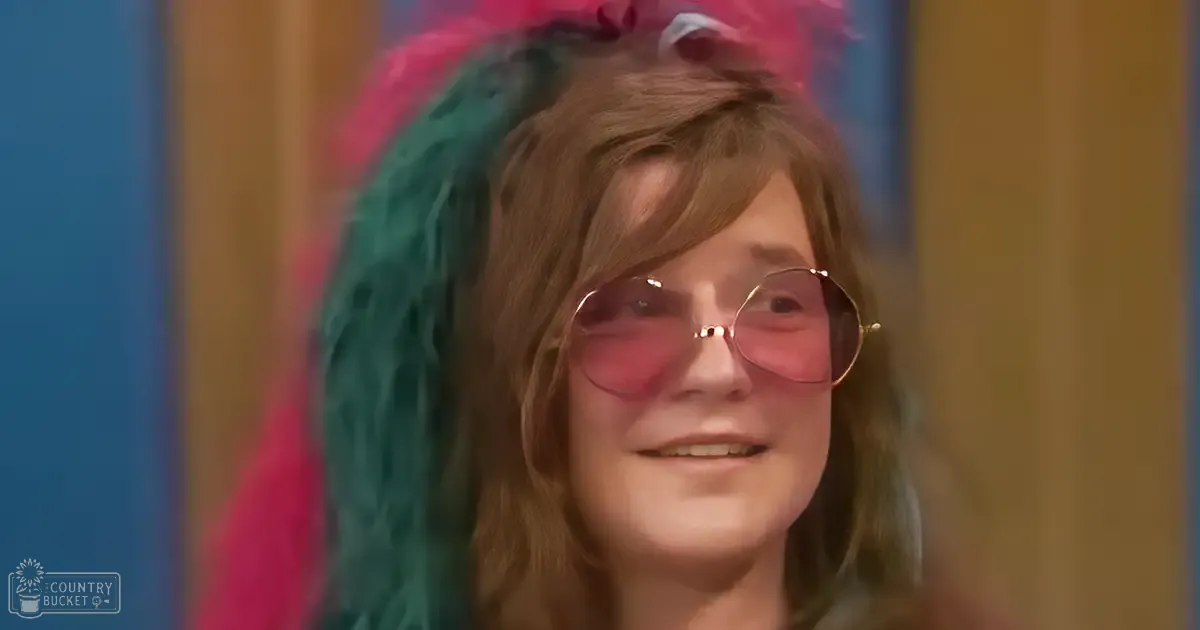 A young adult female wearing round sunglasses and shoulder-length light brown hair in an indoor setting with a blurred wooden panel.
