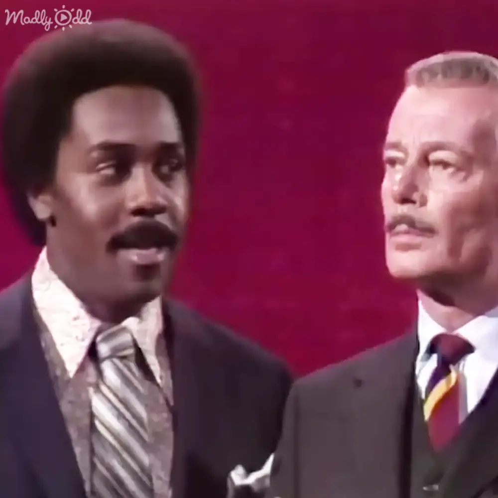 Two middle-aged men dressed in formal attire stand against a solid red backdrop, suggesting an indoor stage setting.