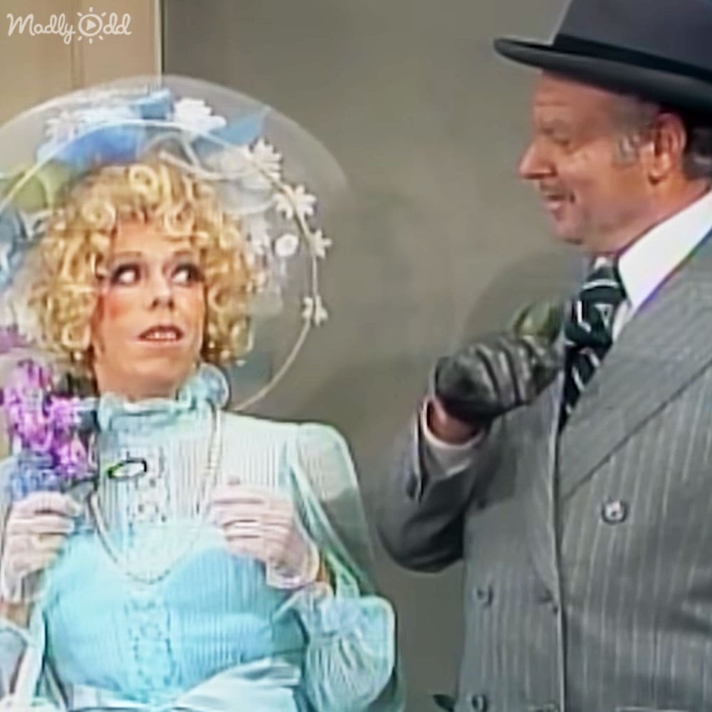 A woman in a vintage light blue dress and white gloves stands indoors alongside an adult male wearing a gray pinstriped suit with a black fedora. The scene suggests a mid-20th-century television show or film setting.