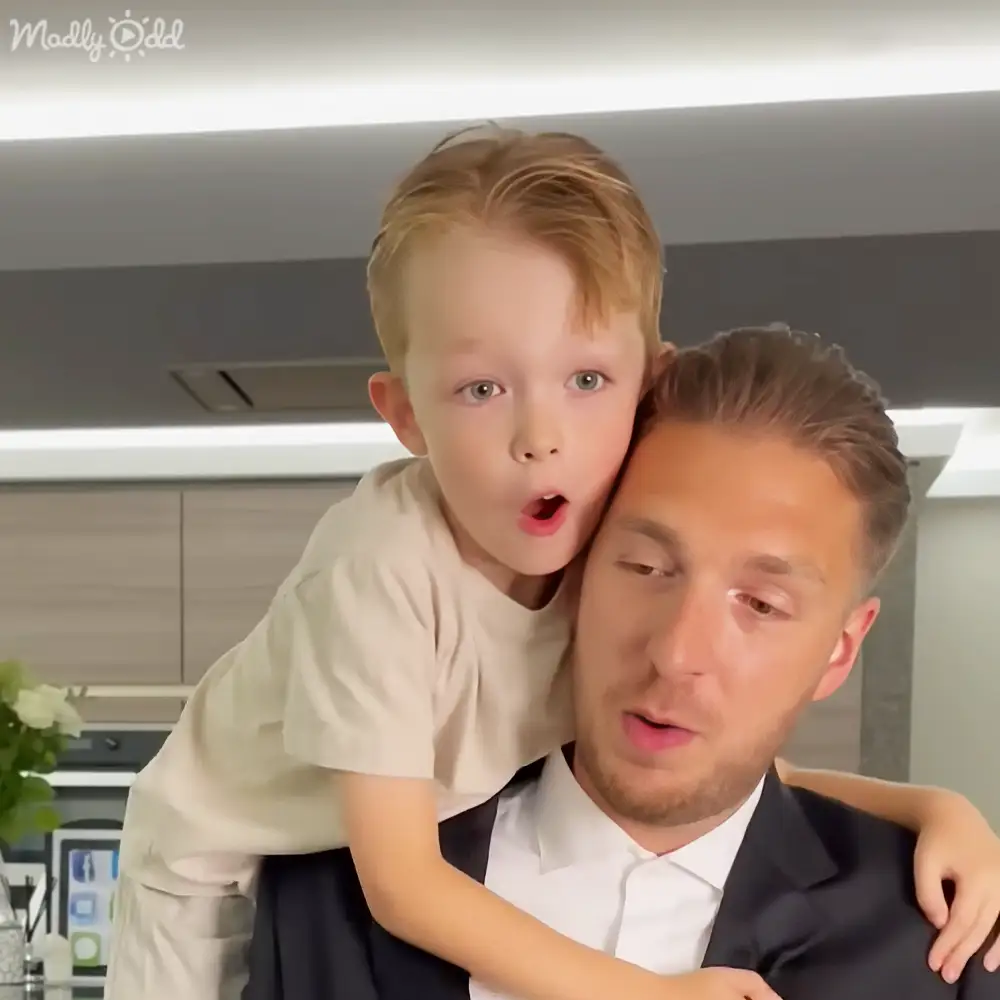 A young boy with short blond hair wearing a cream-colored shirt leans against an adult male in a modern kitchen setting during daytime.