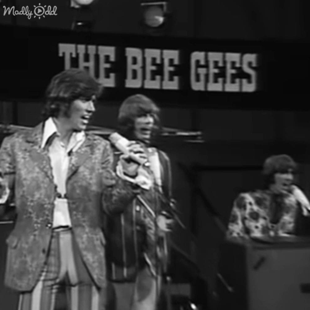 The Bee Gees performing on stage with a microphone stand visible behind them; backdrop features their name.