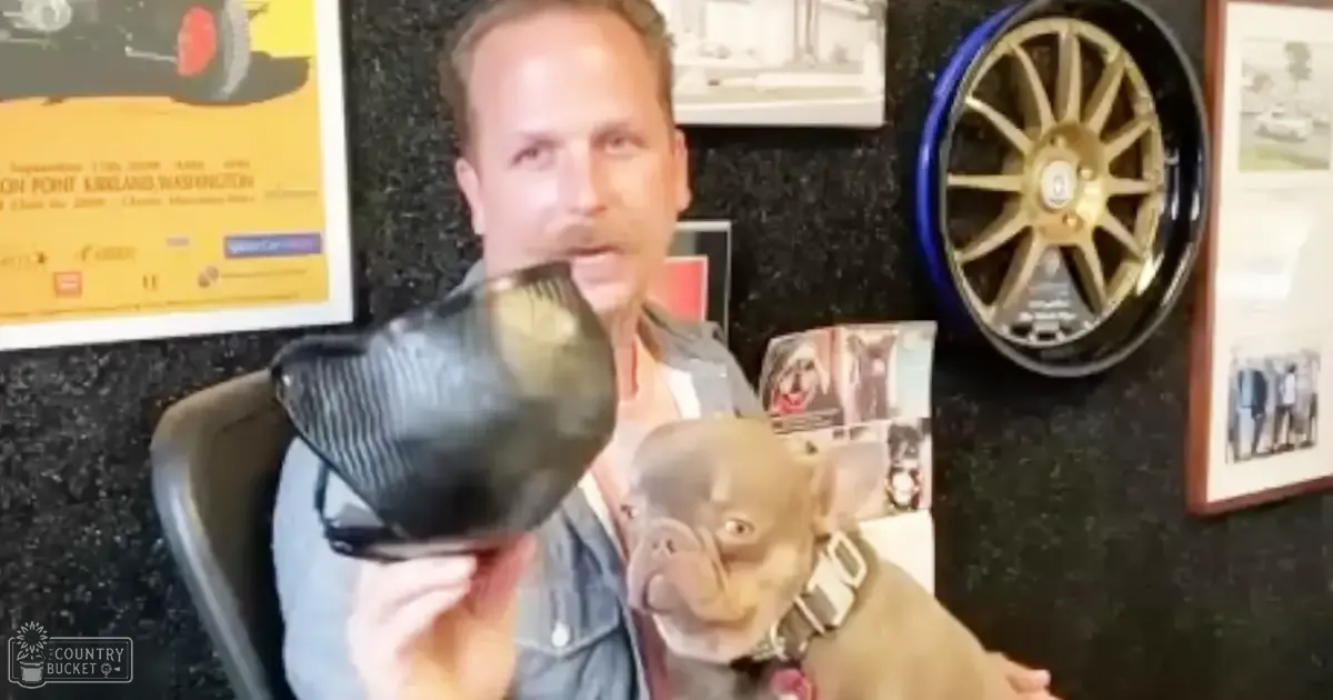 A middle-aged male wearing a denim jacket indoors with a small brown Chihuahua, surrounded by framed items on dark walls suggesting a personal space.