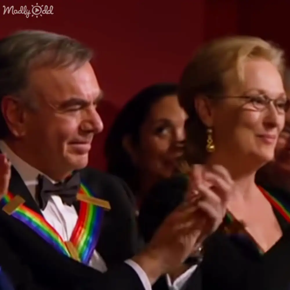 Adult male in a formal event, possibly an award ceremony, wearing a black tuxedo with a colorful medal ribbon pinned to his lapel; adult female in dark formal attire with gold earrings and glasses stands nearby. The setting is indoors at a professional event with red background lighting.