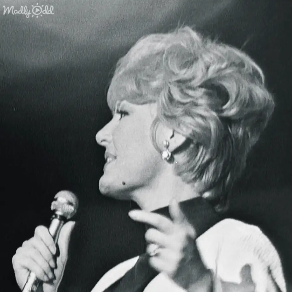 A middle-aged female performer on stage wearing a mid-20th century-style white garment with long sleeves and high neckline, holding a microphone in an indoor venue with minimalistic background lighting.