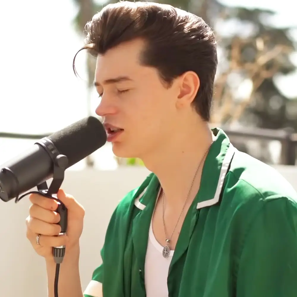 A young adult male wearing a green jacket with white trim, singing into a microphone outdoors.