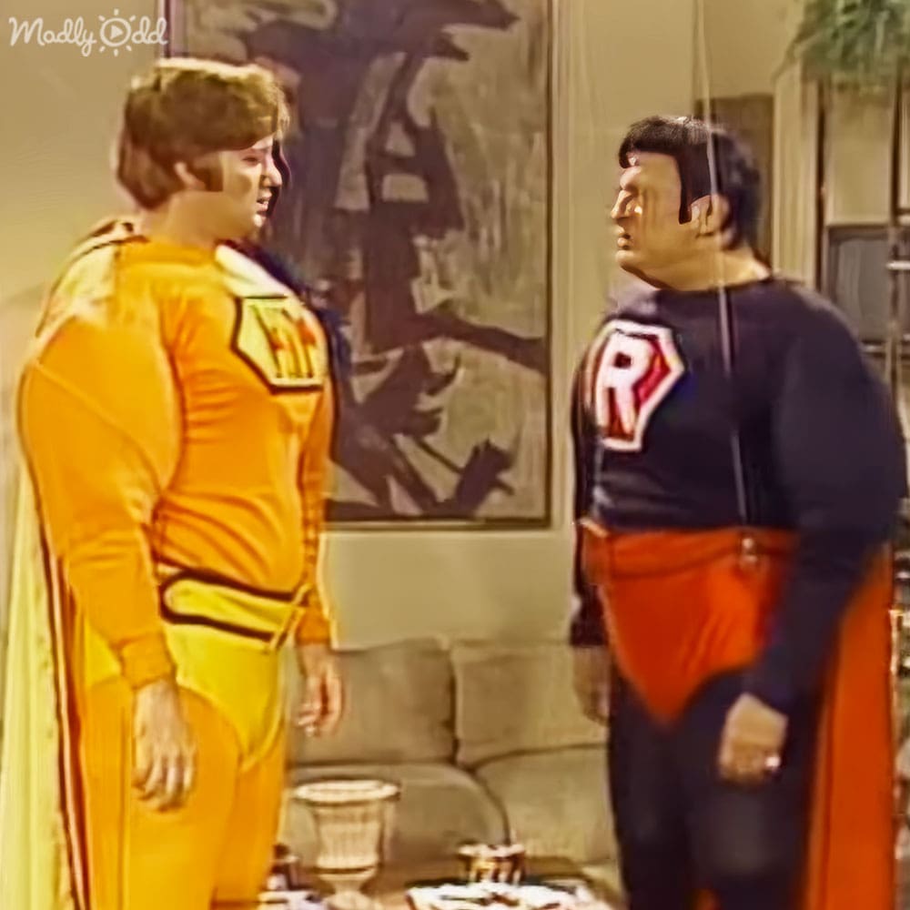 A young adult male in a bright yellow jumpsuit stands beside an adult male wearing a dark blue shirt with an orange sash and 'R' emblem, set against the backdrop of a stone fireplace and a natural scene painting. The indoor environment suggests a living room or studio set designed for filming.