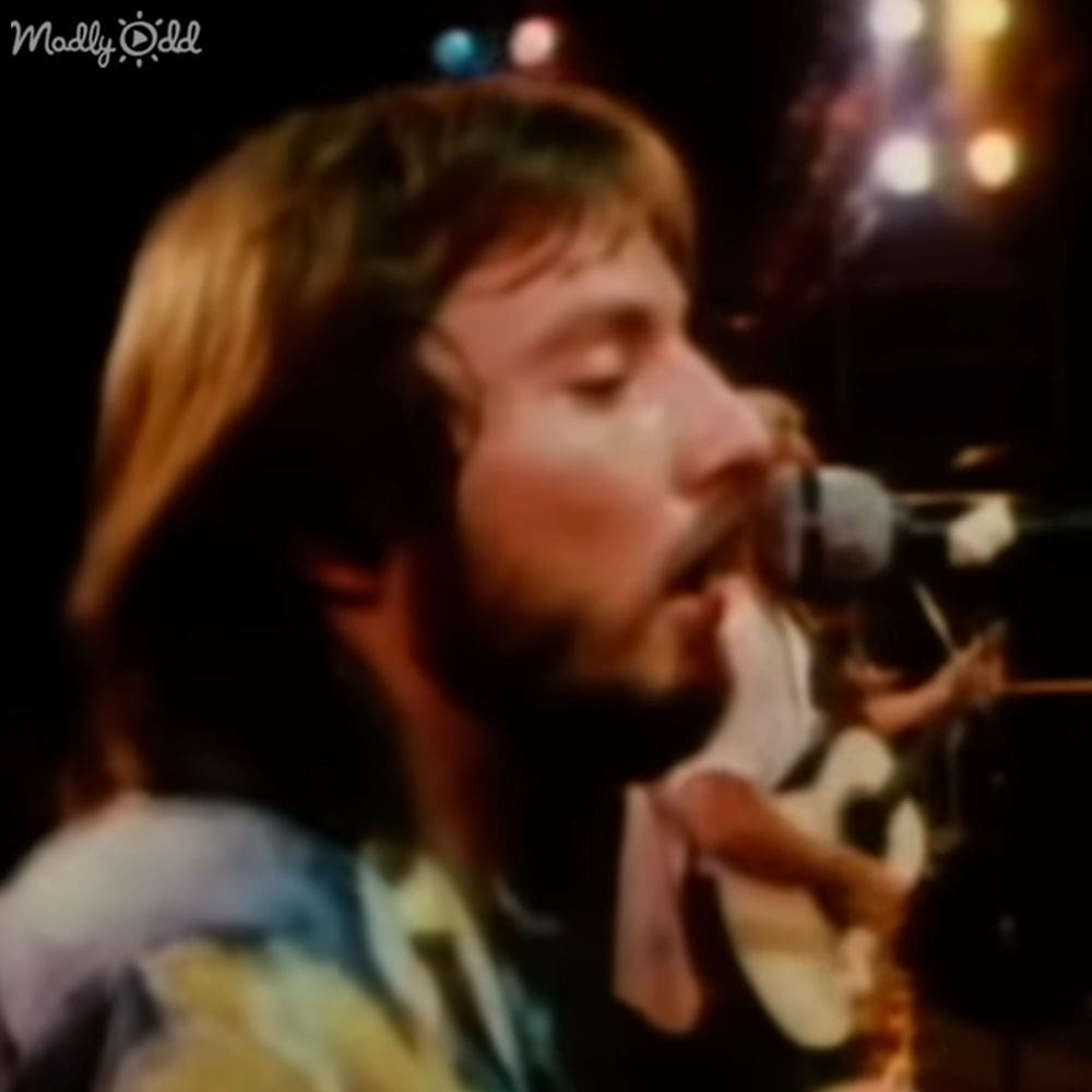 An adult male singer wearing a loose-fitting 1960s or 70s style tie-dye shirt, holding a guitar strap, singing into a microphone at a concert setting with stage lighting and dark background.