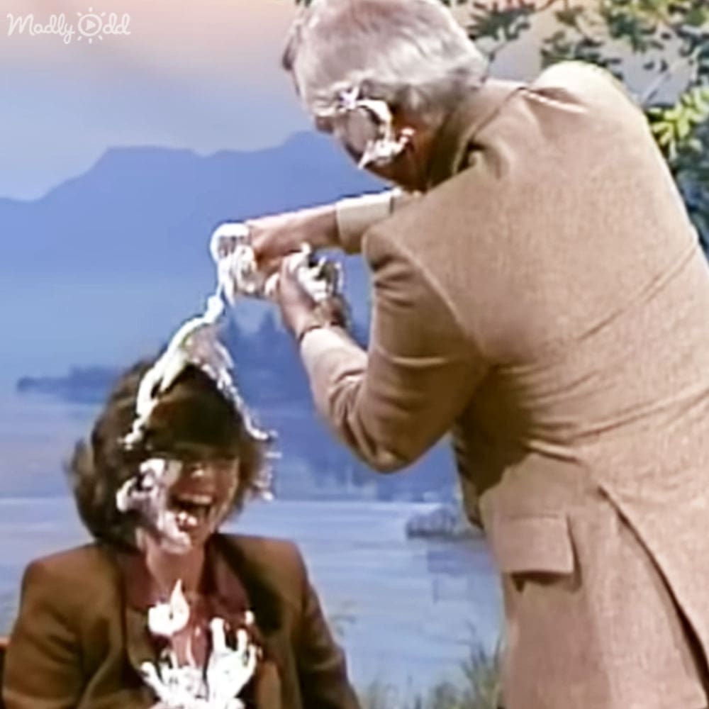 Two middle-aged adults interact humorously near water on a bright spring day in front of mountains.