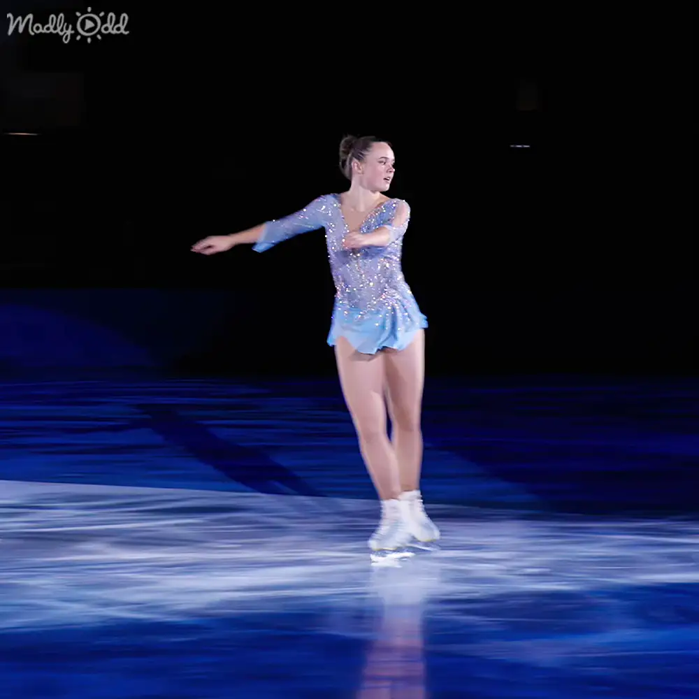 A young adult female figure skater in a light blue, sparkly figure skating costume performs on an indoor ice rink with dark surroundings at night.