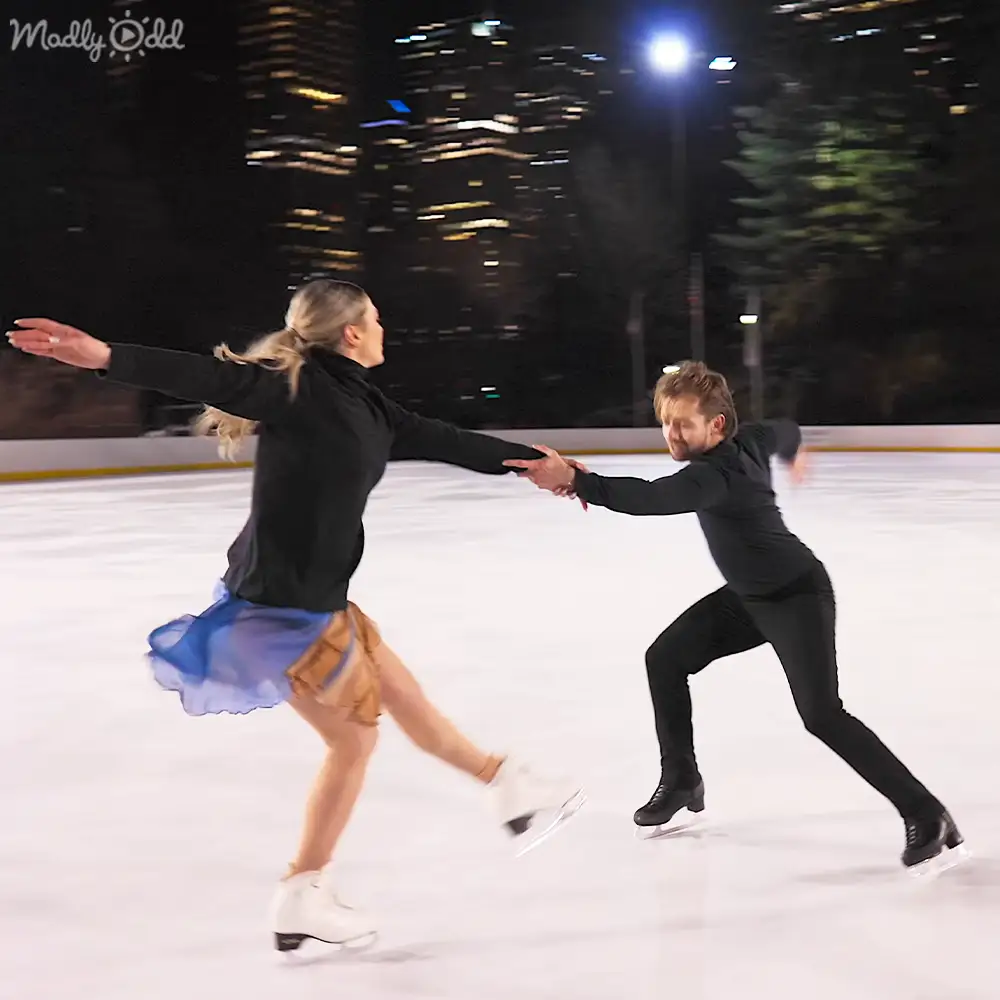 A couple ice skating at an outdoor rink surrounded by a nighttime urban cityscape with illuminated buildings and trees.