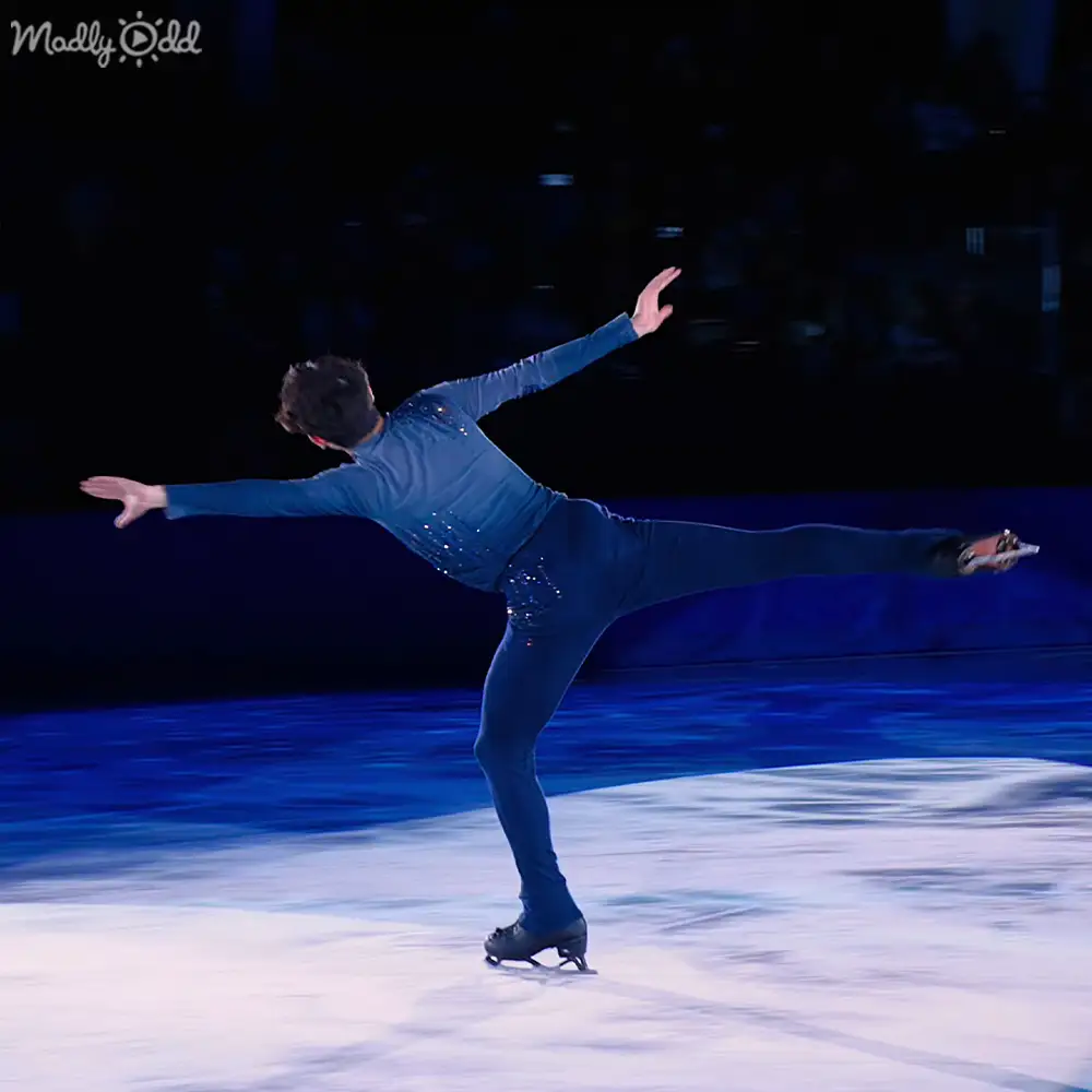 A young adult male figure skater in a blue, form-fitting skating costume with embellishments performs on ice under spotlighting within an indoor arena.