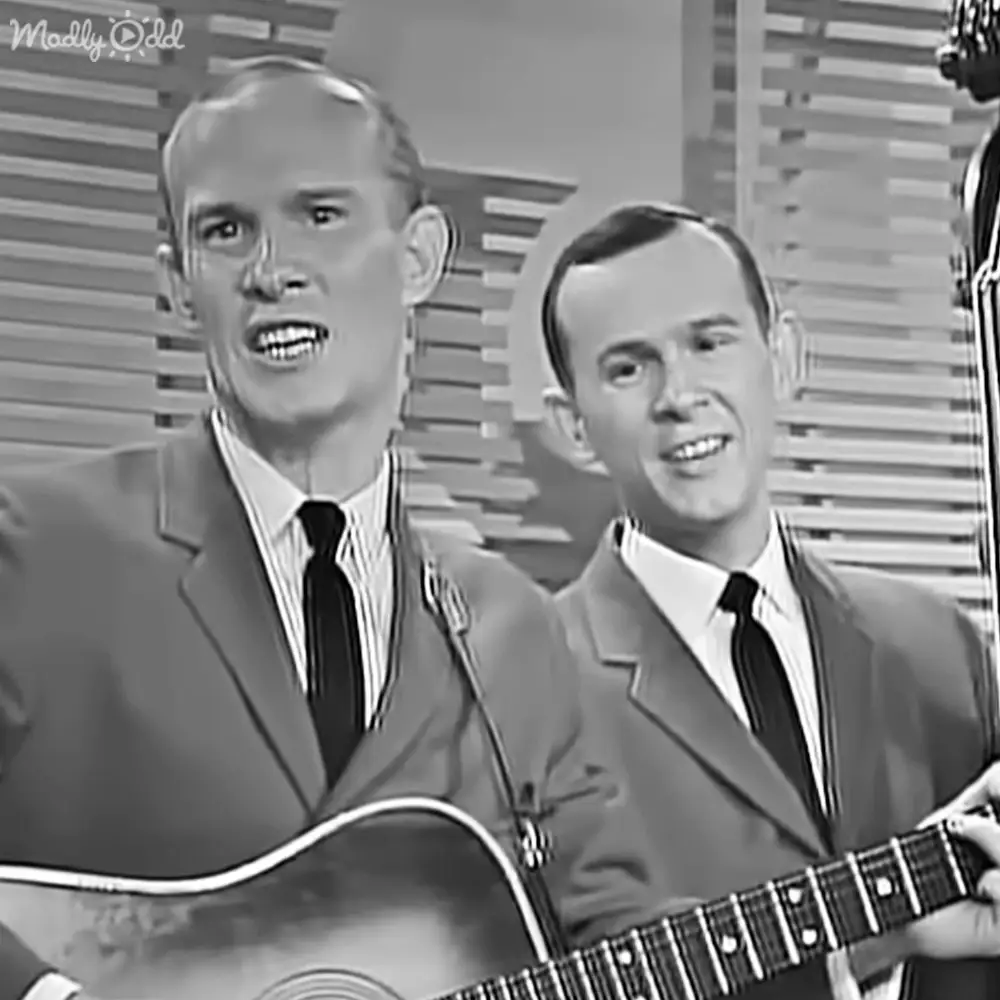 Two adult males in a mid-20th century style suit performing on stage with horizontal blinds as the backdrop.