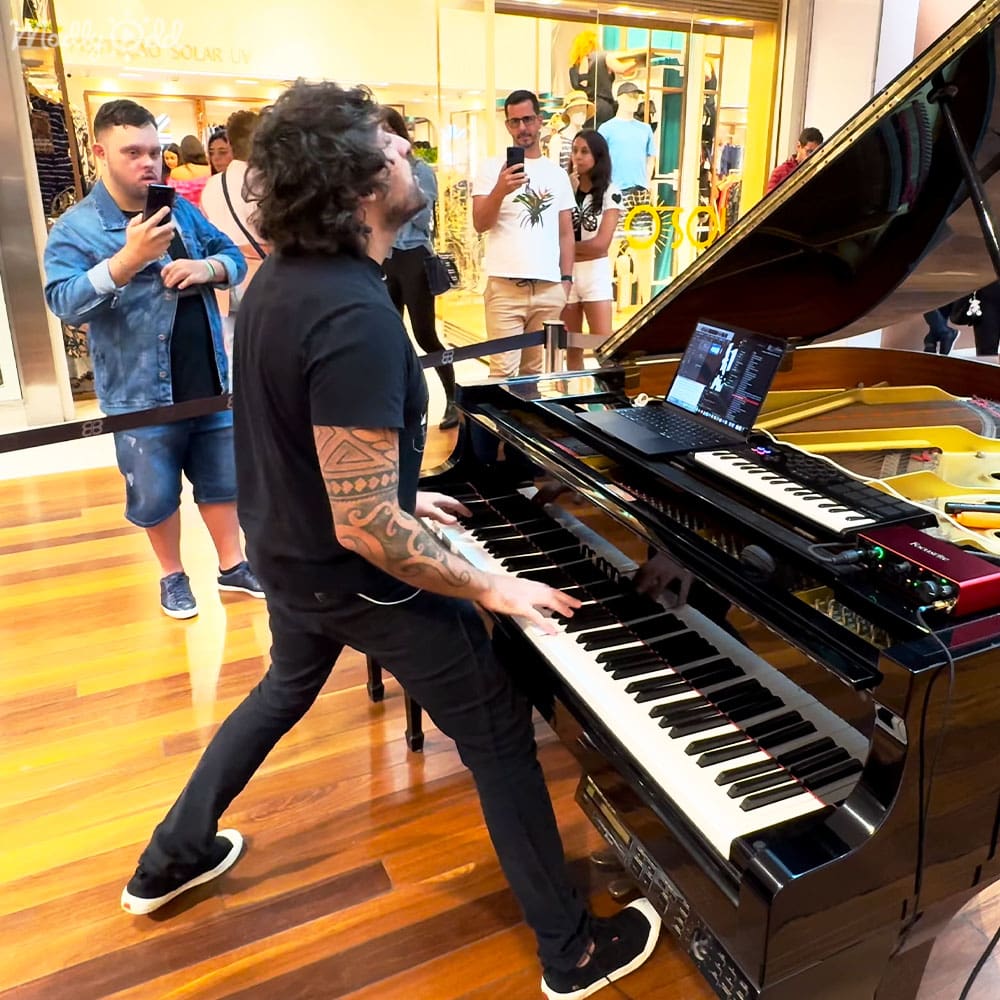 A young adult male pianist in a black t-shirt and pants plays a grand piano with a laptop connected to it, surrounded by other individuals holding smartphones inside a shopping mall.
