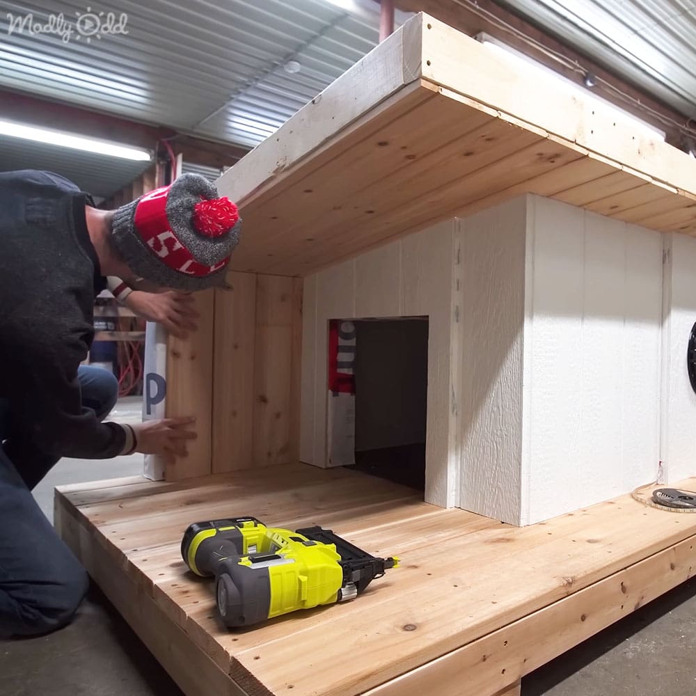 An adult male in a workshop setting, engaged in woodworking activities wearing a dark jacket with long sleeves, red and gray beanie, and blue jeans.