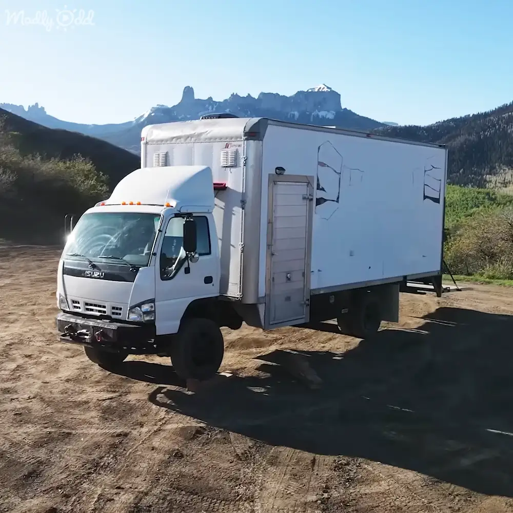 White Isuzu truck parked in rural area with mountains, clear sky, late spring/summer, mid-morning or early afternoon light.