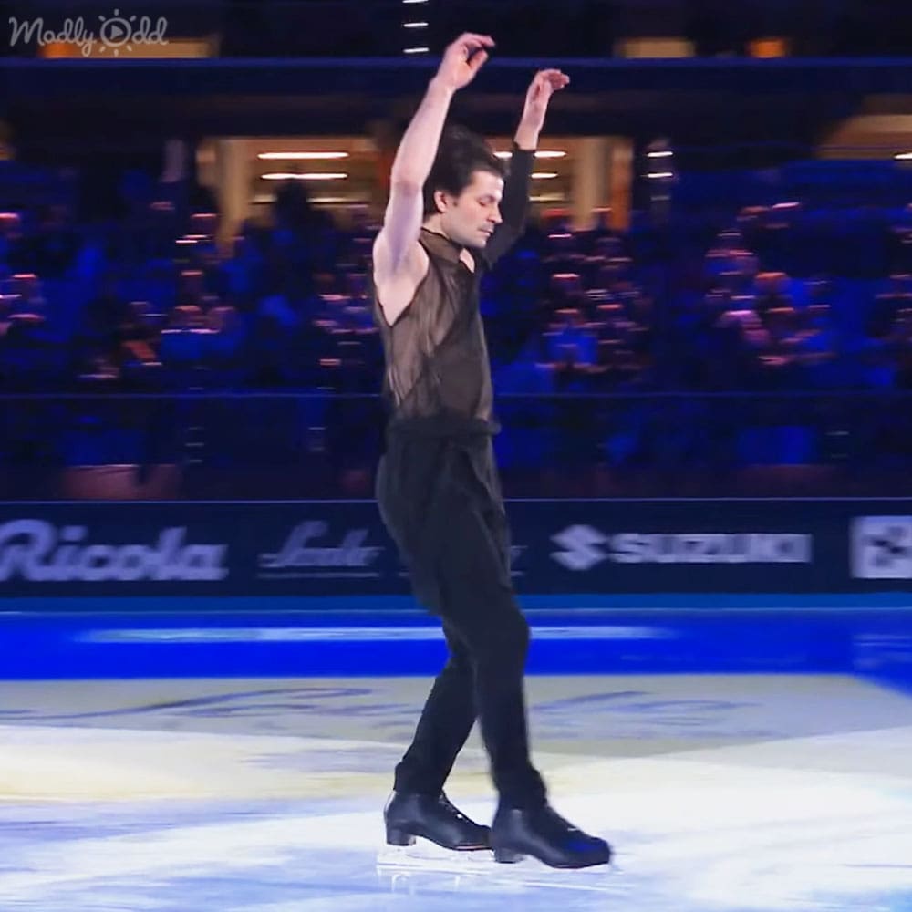 An adult male figure skater performs at an indoor ice rink with blue lighting, surrounded by seated spectators and promotional signage.