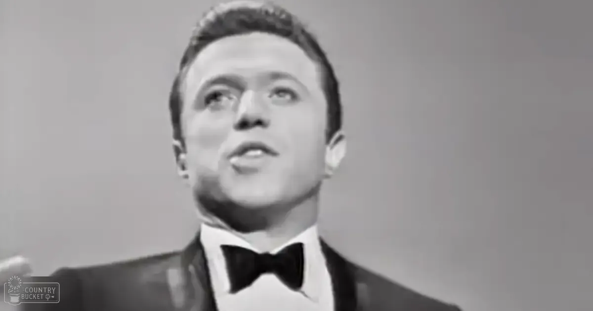 A young adult male in a fitted mid-20th-century tuxedo with a bow tie, standing indoors under natural light.