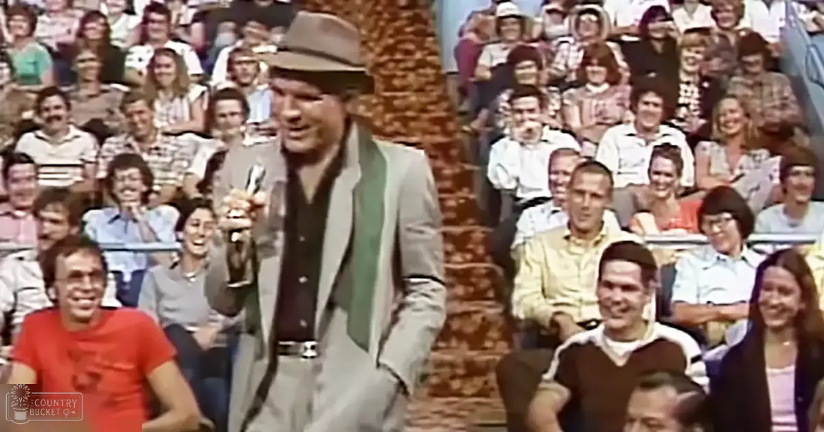 A middle-aged man in a light-colored suit jacket, dark shirt, and fedora-style hat holding a microphone on stage in an indoor entertainment setting with an audience of various ages seated in rows.