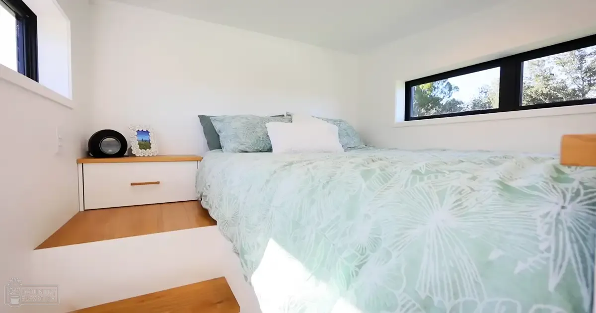 A modern bedroom with a bed, two nightstands, framed picture, clock, windows showing greenery, white walls, light green floral duvet cover, and brown nightstands.