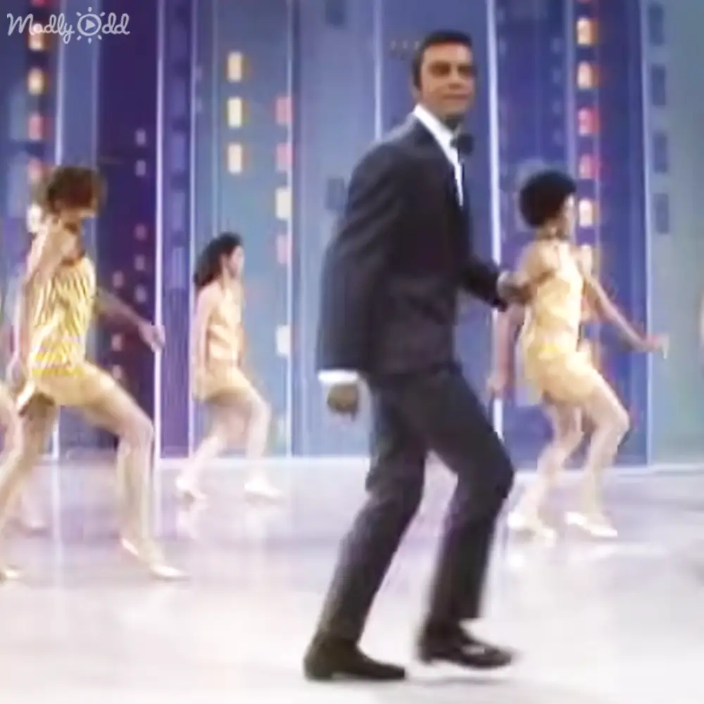 A man in a mid-20th century-style dark suit dances amidst young children wearing matching gold dresses in an indoor studio setting with vertical panels and small lights or windows.