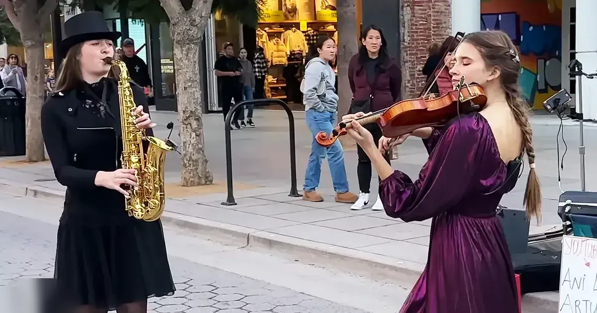 A street performance scene with young adult female saxophonist, another playing violin, and onlookers under trees near storefronts; spring or summer daytime setting.