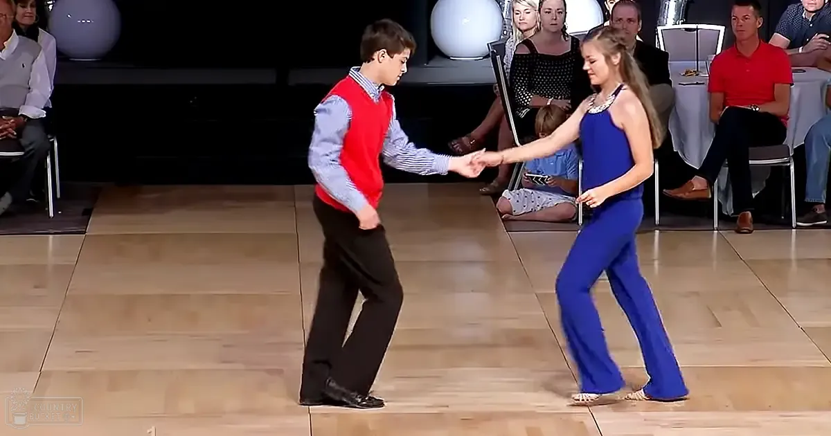 A dance pair performing in a studio setting with chairs for spectators, tables with white tablecloths, and decorative spherical objects on stands behind them.